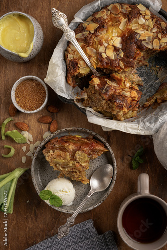 Top view of baked English rhubarb pudding slice with ice cream, mint and tea cup, vertical flat lay of traditional custard cake in skillet on wooden background