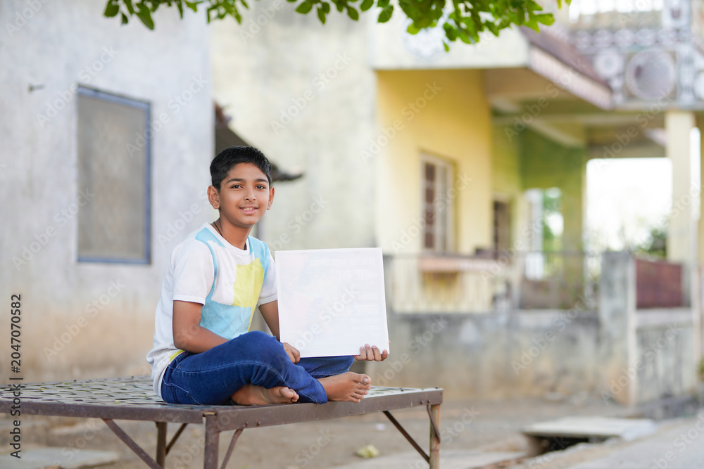 Fototapeta premium Indian schoolboy sitting outdoors in a rural village while holding and pointing at a blank display board