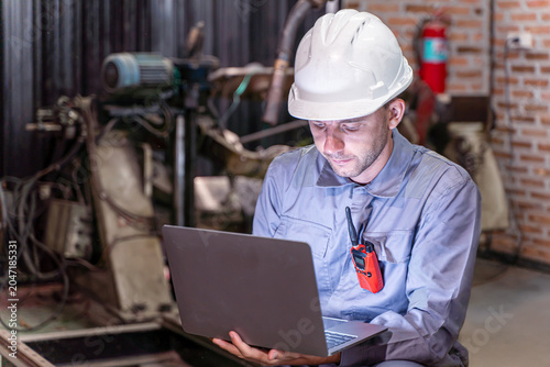White male engineer in safety gear inspects production data on a laptop inside an industrial