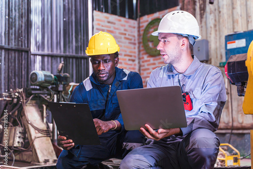 White man engineer talking with his friend black man engineer in hard hats discuss new project while using laptop. They make showing gestures in an industrial workshop.