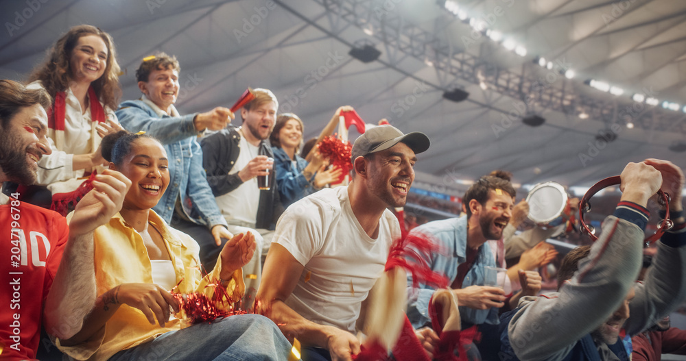 Naklejka premium Diverse Fans at a Soccer Football Championship Laugh, Shout and Clap in Stadium Seating After a Crucial Moment, Waving Red Scarves and a Tambourine as Relief and Joy Spread Across Their Faces.