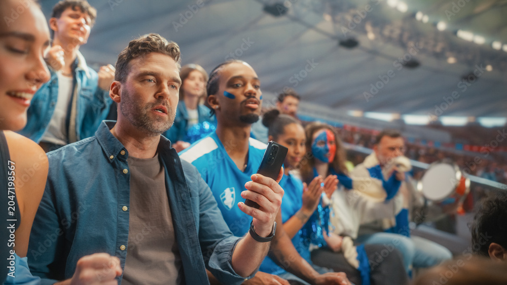 Obraz premium Portrait of a White Male Fan Checking a Smartphone in Stadium Stands at a Soccer Football Championship Tournament, Surrounded by a Happy Excited Diverse Crowd of Smiling Supporters Cheering a Goal.
