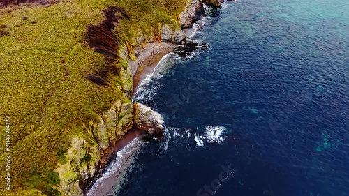 Elevated side perspective of high rocky coast meeting dark shoreline and deep blue ocean. Drone shot of steep grassy cliffs and secluded beach.