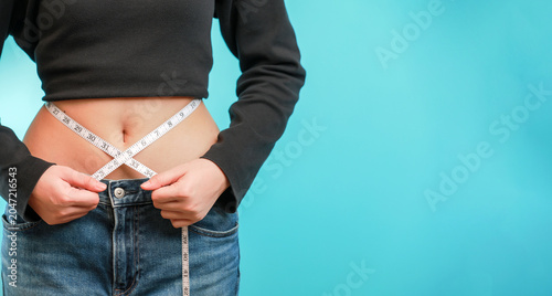 Confident woman measures slim waist with white tape during successful healthy weight loss journey against plain blue background