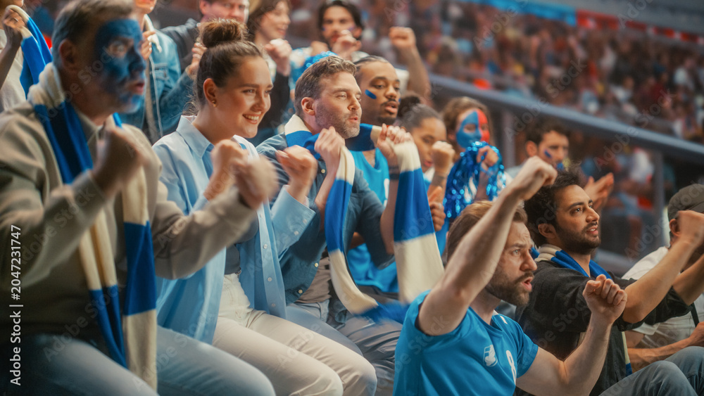 Naklejka premium Diverse Crowd of Excited Male and Female Fans With Painted Faces and Blue Scarves Watching a Tense Soccer Football Championship Match in Stadium Stands, Happy Smiling Supporters Cheer for Their Team.