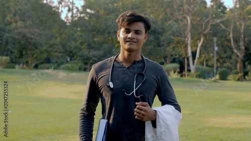 Young aspiring male doctor with a stethoscope on the neck standing in the park. Medium shot of a charming studious boy with a stethoscope  a doctor's overcoat  and a book in an open area
