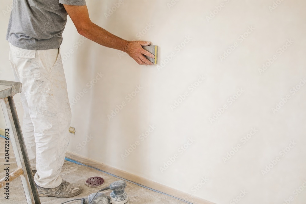© Studio - Professional male painter sanding plaster wall with hand block during home renovation in bright indoor room, construction worker preparing surface for painting, copy space