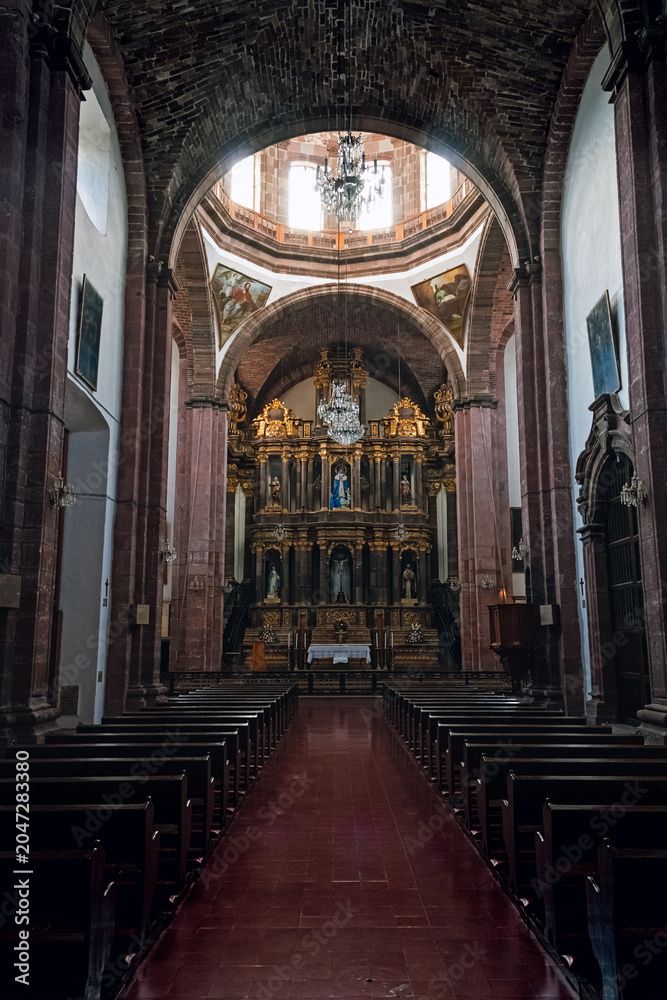 Obraz premium Grand cathedral interior with tall arches and golden altar. Dark pews line a red-laid floor leading to sacred space. Chandelier hangs above, casting soft glow
