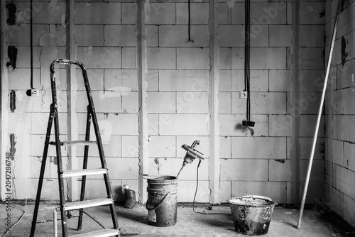 Interior view of room during renovation. Bare block wall with exposed wiring and wall leveling guides. Blue stepladder, buckets, and electric mixer for plaster black and white photo