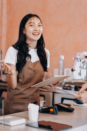 Friendly Asian female barista receives a credit card from a customer for payment. Both women are smiling in a warmly lit coffee shop with a rustic interior. Cashless payments. Small family business