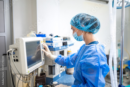Female anesthesiologist adjusting anesthesia machine monitor screen in operating room, healthcare concept. Woman doctor checking patient vital signs on medical display equipment during surgery.