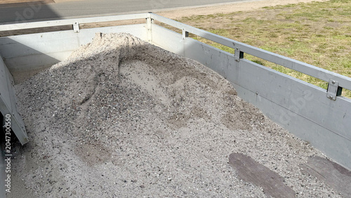 Pile of gravel and sand in the back of a truck bed on a construction site with a grassy area and road visible in the background