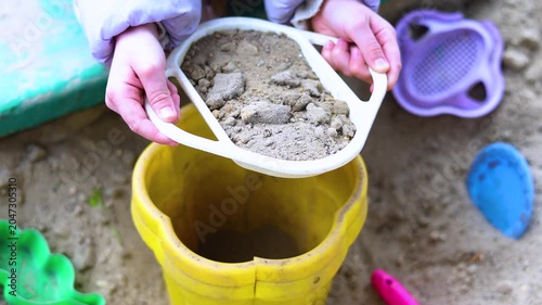 Experience the magic of childhood in this delightful scene where little hands enthusiastically sift and pour sand into a bucket. The carefree spirit of play permeates every moment.