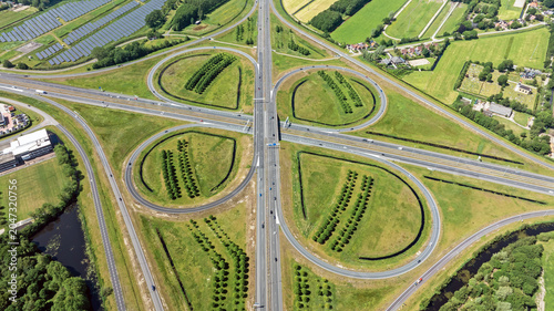 Aerial view of a cloverleaf highway interchange with green fields, solar panels, and surrounding vegetation in Heerenveen, Friesland, Netherlands.