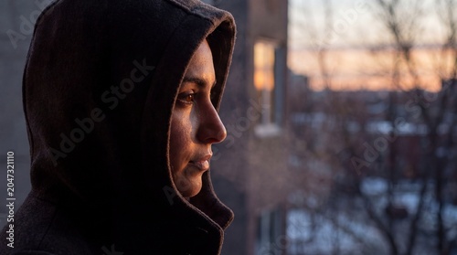 A young woman in a dark hoodie gazes out of a window, her face illuminated by the soft, golden light of dusk, evoking a sense of introspection and solitude.