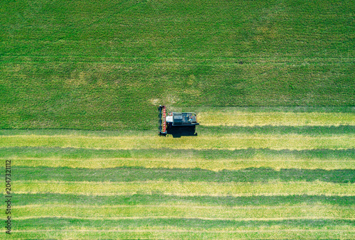 Aerial view captures powerful combine harvester gathers crops on a vast green field under a clear blue sky during a sunny day near