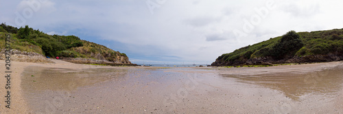 Calm panoramic view of a sandy beach cove at low tide, framed by rocky cliffs and green headlands with sailboats on the distant horizon under an overcast sky.