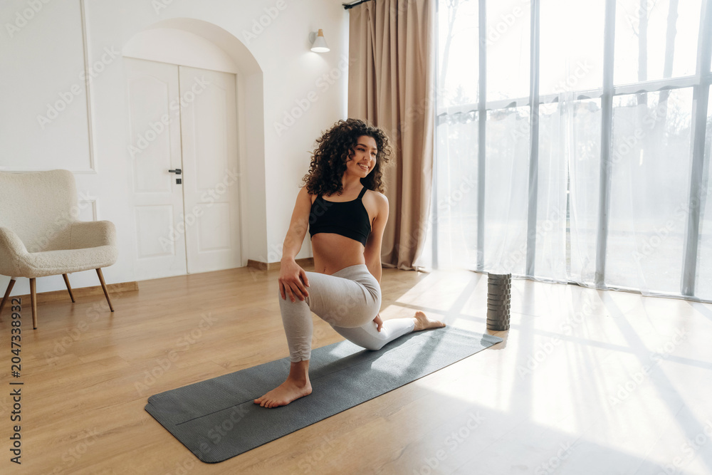 Naklejka premium Female fitness enthusiast with curly hair performs a lunging stretch on a yoga mat in a bright room with large windows and wooden flooring