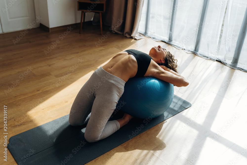 Fototapeta premium Female fitness enthusiast performing an exercise on a blue stability ball in a bright indoor space with wooden flooring and large windows