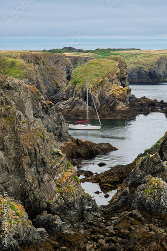 A white sailboat rests in a sheltered rocky cove beneath rugged cliffs and grassy headlands, evoking calm, adventure and coastal solitude under an overcast sky.