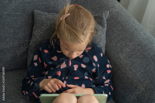 Vászonkép A schoolgirl sits on a grey sofa absorbed in her gadget, staring intently at the screen, highlighting concerns about digital addiction in children