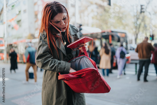 Quadro su tela A young woman talks on her phone while searching through a red backpack on a city sidewalk