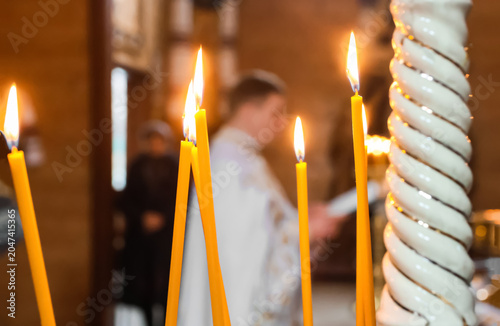 Close-up of burning yellow wax candles inside an orthodox church. Defocused priest reading a holy book in the background during a religious ceremony or liturgy
