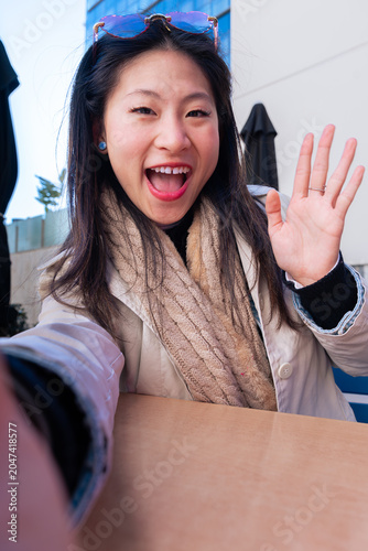 Vertical shot of a smiling young woman waving her hand to say hello while taking a selfie or being on a video call, wearing stylish red sunglasses, captured in a bright outdoor shopping mall area