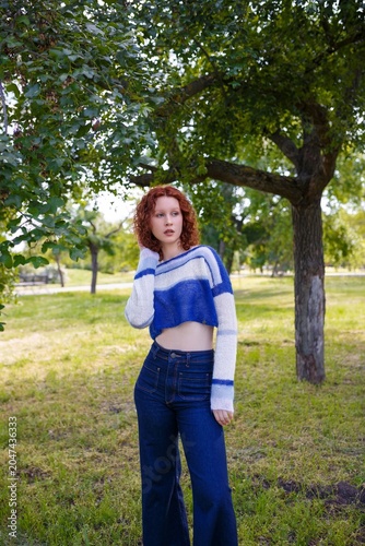 Redhead teenage girl in jeans and blue striped knit crop sweater posing on green summer background