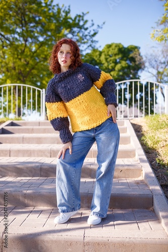 Redhead teenage girl model in chunky knit sweater and jeans on stairs background. Bulky knitwear, fashion and style concept