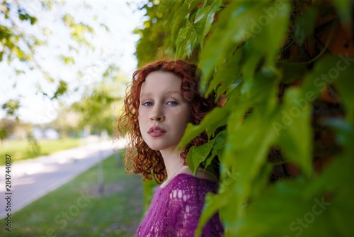Redhead teenage girl in knitted purple transparent sweater posing at green ivy wall. Close up portrait. Clothing and fashion concept