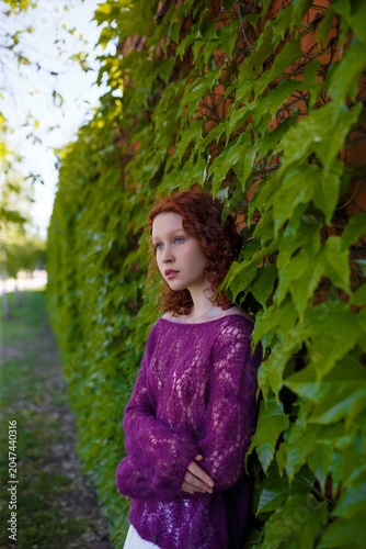 Redhead teenage girl in knitted purple transparent sweater posing at green ivy wall. Clothing and fashion concept