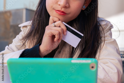 Thoughtful East Asian young woman holding a blank white credit card while looking at a tablet screen. She sits at an outdoor urban cafe patio, considering an online purchase, lifestyle shopping