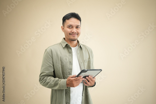 Young Asian man holding a tablet with a warm confident smile, facing slightly sideways in casual olive green shirt against beige studio background