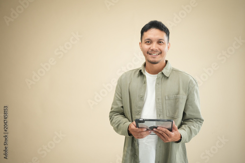 Young Asian man holding a tablet with both hands and smiling broadly at camera, wearing casual olive green shirt against beige studio background