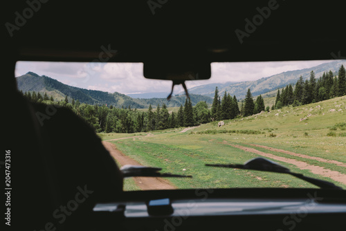 View from inside a vehicle of a winding dirt road across green meadows and pine forest in Ak-Kem Valley, Altai Republic, Russia, under a cloudy summer sky