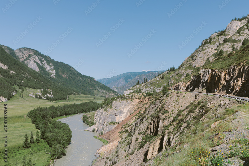 Fototapeta premium Wide summer landscape of Chuysky Trakt highway carved into rocky cliffs above the Katun River, with green valley and mountains, Altai Republic, Russia