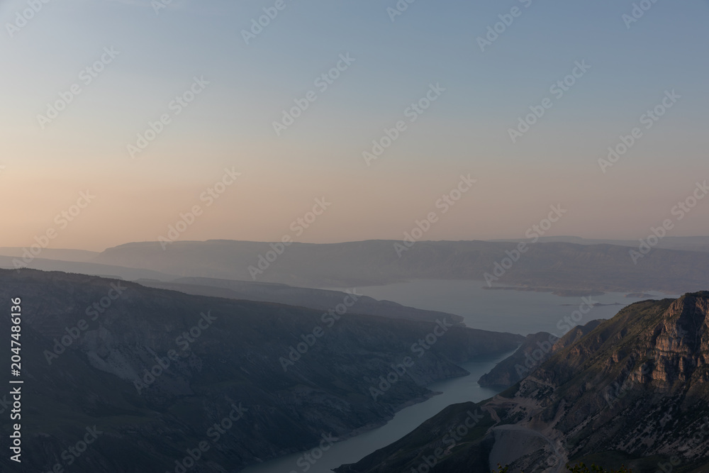 Fototapeta premium Wide panoramic landscape of Sulak Canyon with winding river and layered mountain ridges at sunrise, atmospheric haze and calm sky in Dagestan, Russia
