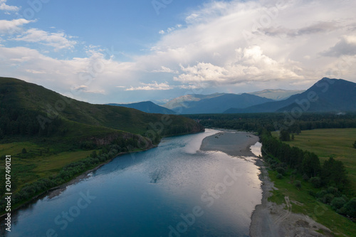 Aerial landscape of a winding river through forest and meadow with distant mountain ridges under dramatic clouds near Zamulta, Altai Republic, Russia at dusk