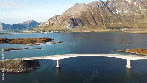 A bridge connects islands in Norway as boats move slowly in the water. Clouds float above and hills rise in the distance. This view shows the landscape and water.