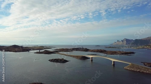 A bridge connects islands in Norway as boats move slowly in the water. Clouds float above and hills rise in the distance. This view shows the landscape and water.
