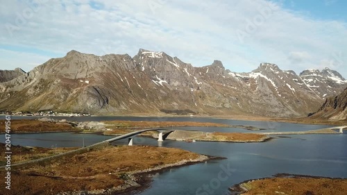A bridge connects islands in Norway as boats move slowly in the water. Clouds float above and hills rise in the distance. This view shows the landscape and water.