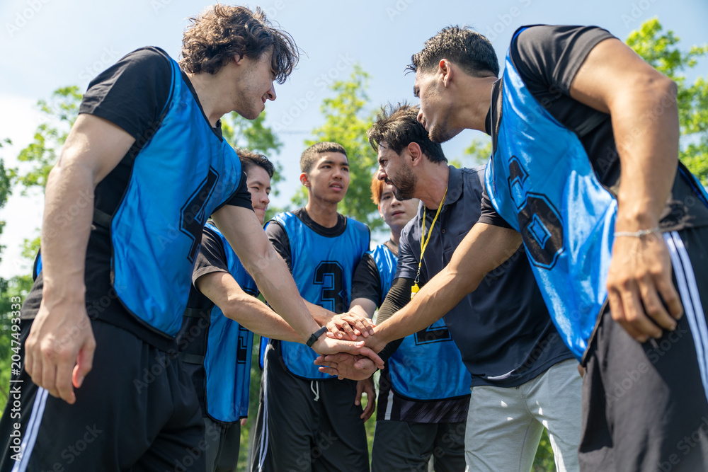 Naklejka premium Diverse group of young soccer players stacking hands with coach before football match outdoor. Multiethnic athletes huddle together showing team unity at green field