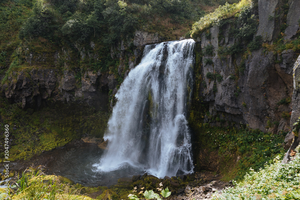 Fototapeta premium Powerful waterfall pours over a basalt cliff into a misty plunge pool surrounded by mossy rocks and green forest vegetation, tranquil nature scenery outdoors