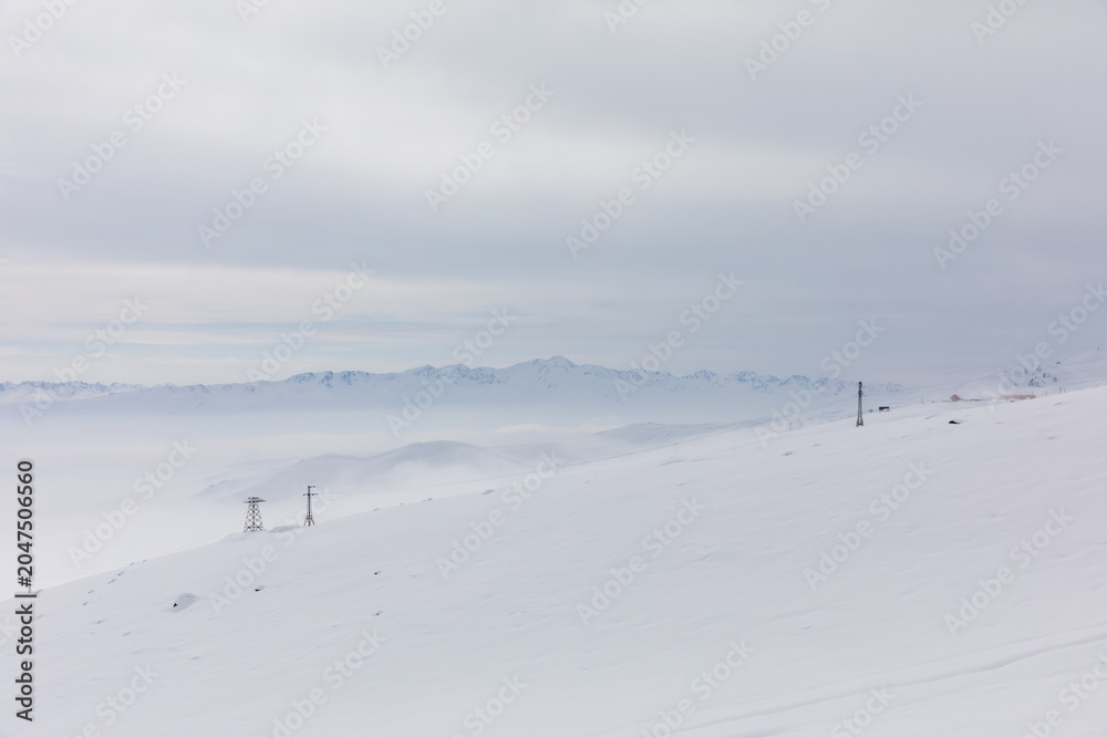 Fototapeta premium Wide winter view of Ala-Bel Pass with fresh snowdrifts, low fog and distant mountain ridge under an overcast sky, Kyrgyzstan, Central Asia travel scenery