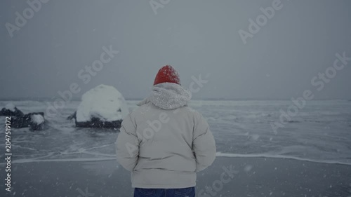 Person in white puffy coat and red hat standing on snowy beach facing grey sea