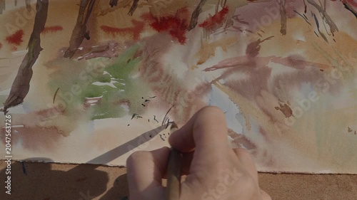 Close-up, the artist's hand paints a watercolor landscape on a sunny day in the forest