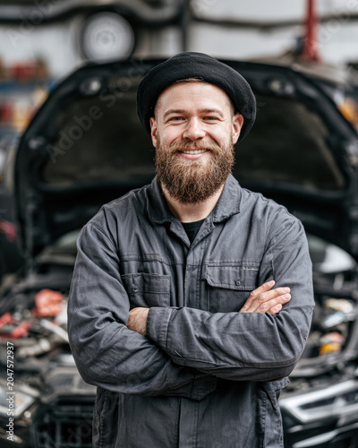 Bearded mechanic in a hat smiles confidently with arms crossed in front of an open car hood in a garage workshop.