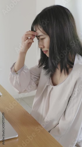 Asian woman holding her head with a troubled expression while working on laptop