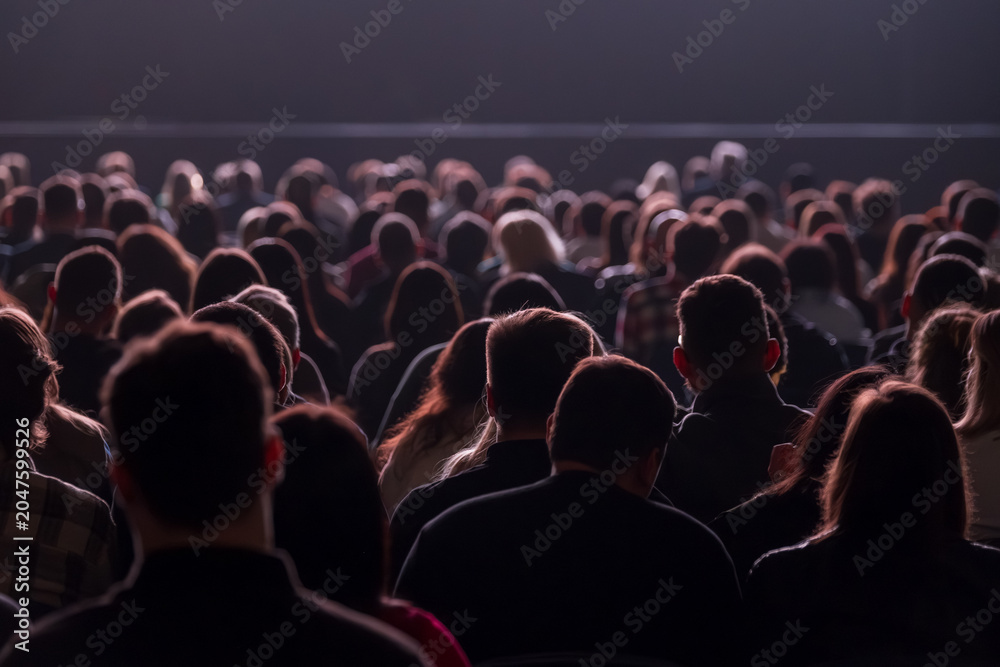 Obraz premium Large seated audience watching performance in dim auditorium, silhouettes and backlit heads create quiet, focused atmosphere.